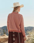 A woman in The Great Inc.'s The Varsity Cardigan stands outdoors with her back to the camera, facing desert hills and cacti under a clear sky.