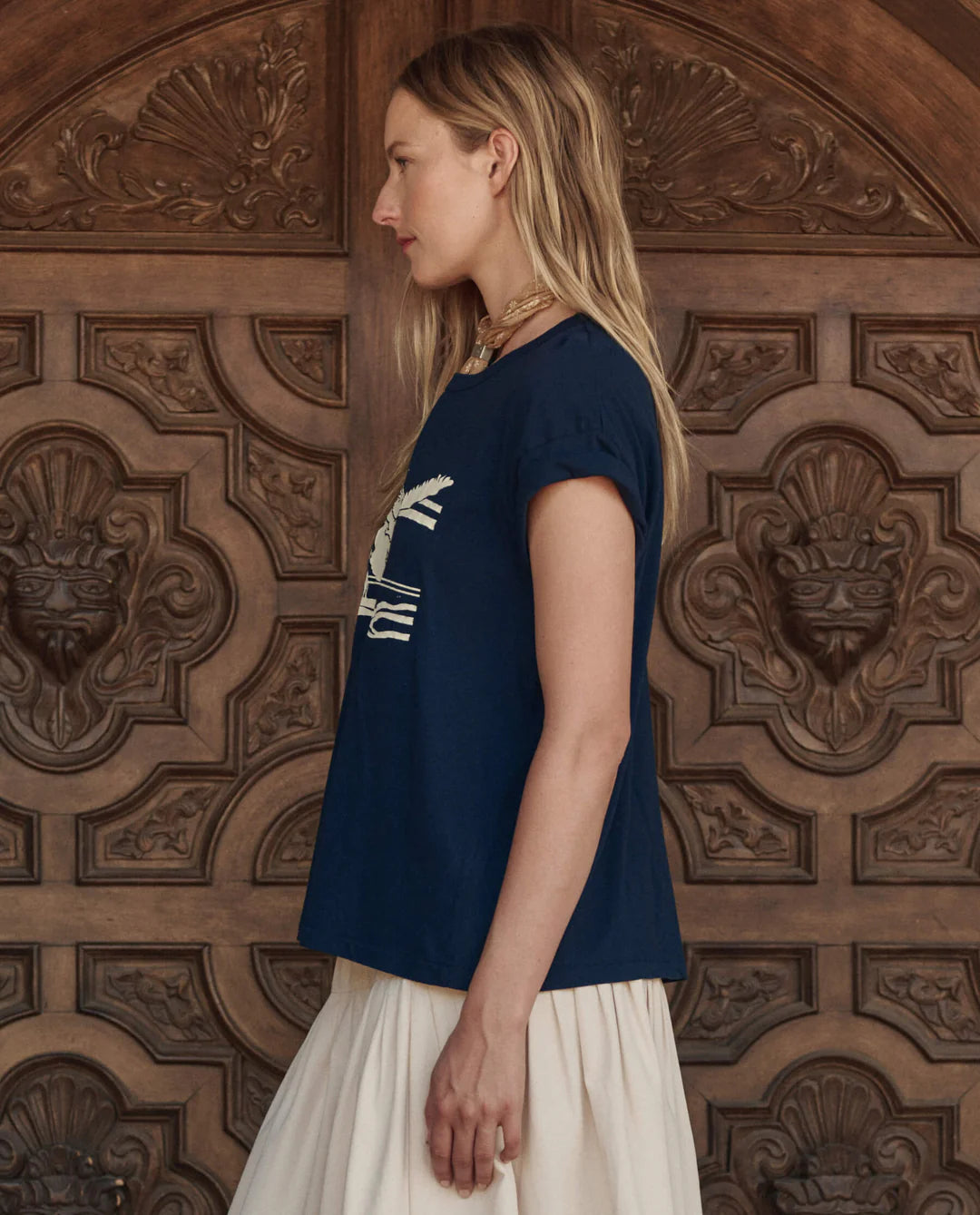 A woman with long blonde hair, wearing The Great Inc.'s The Boxy Crew w/ Everglade Graphic in navy blue and a cream skirt, stands in profile against an ornate carved wooden door.