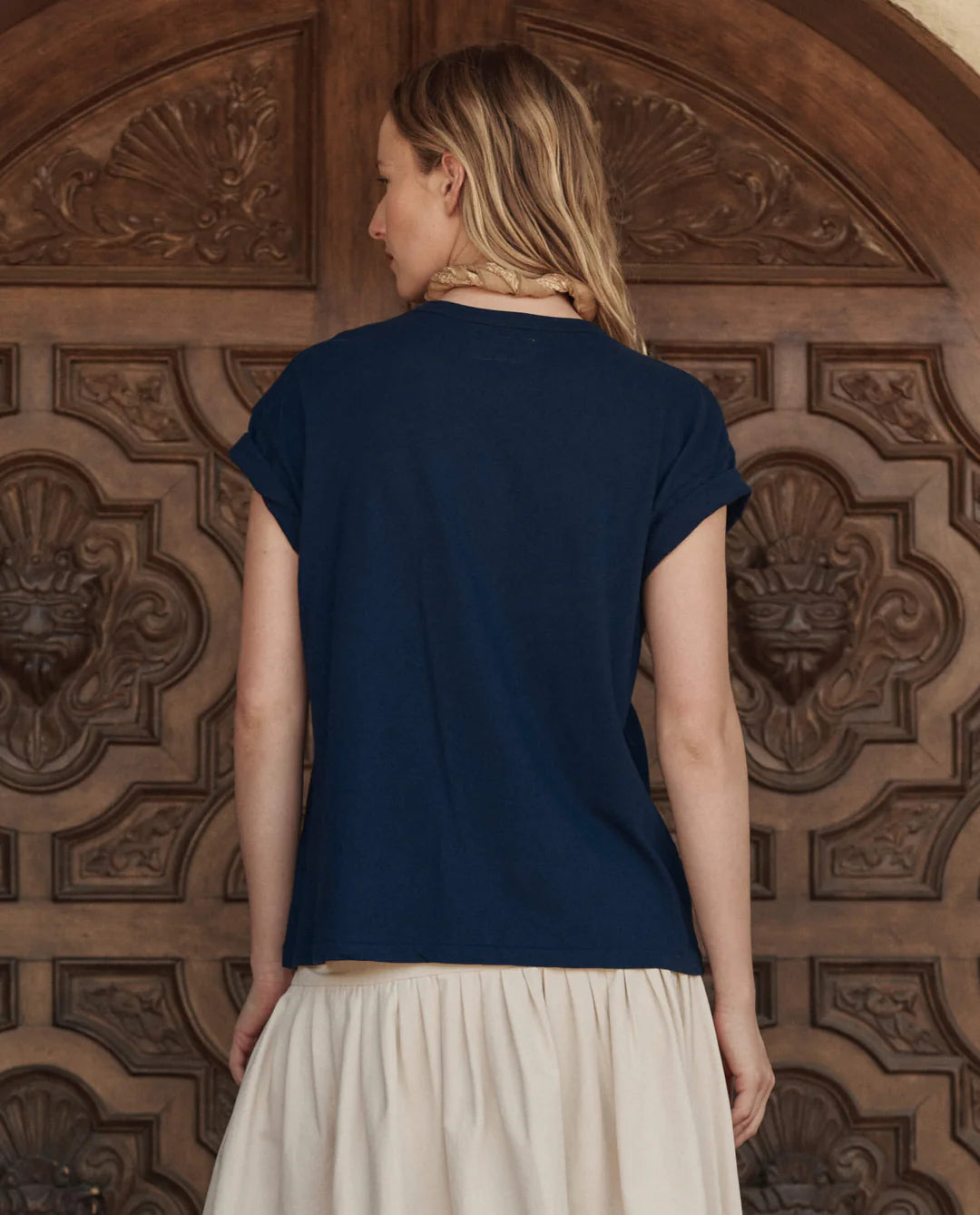 A blonde woman stands with her back to the camera in front of an ornate Los Angeles wooden door, wearing The Boxy Crew w/ Everglade Graphic by The Great Inc. and a cream skirt.