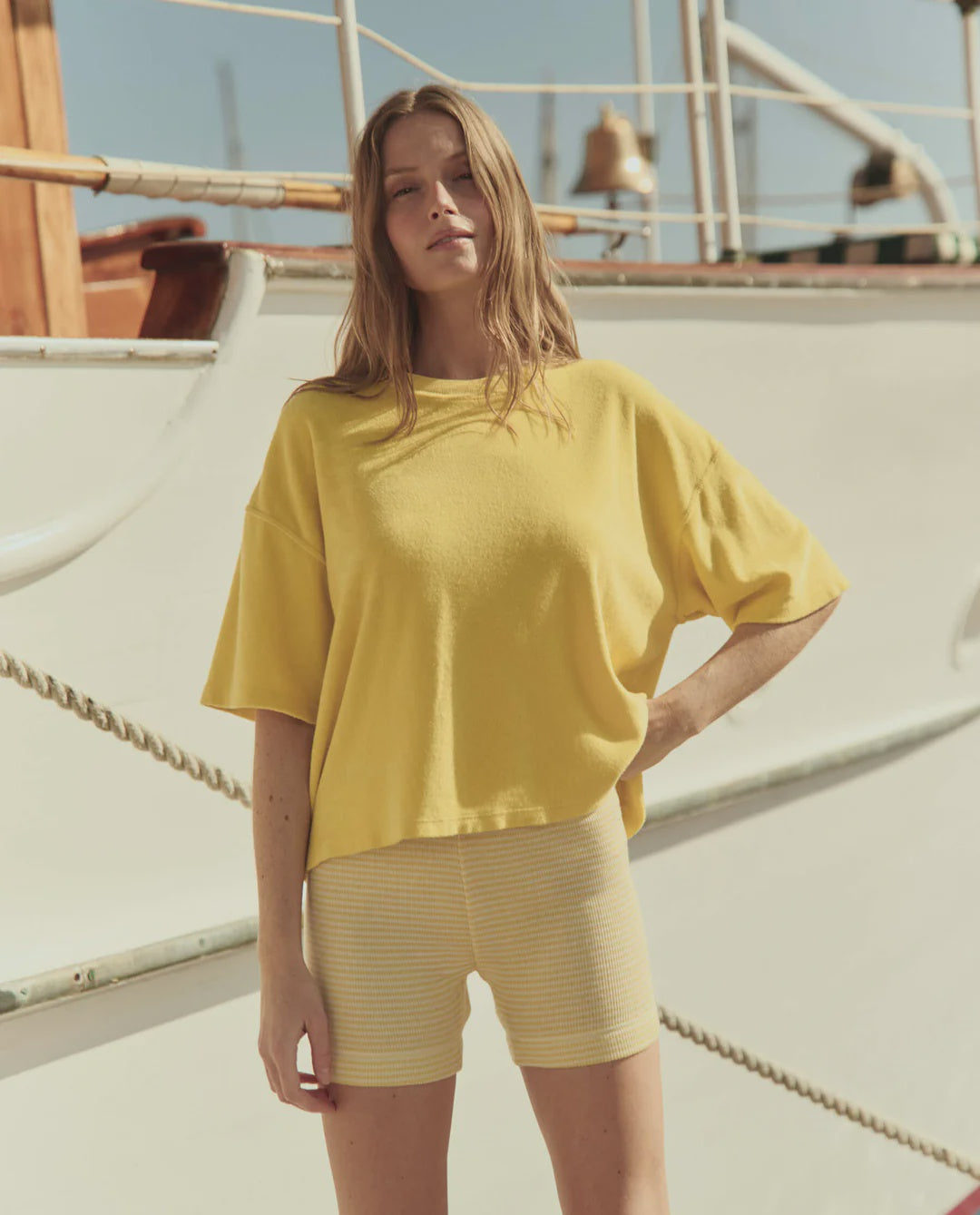 A woman stands outdoors by a boat, wearing The Micro Terry Track Crew by The Great Inc., a loose yellow T-shirt with an oversized silhouette, and yellow striped shorts. Her long light brown hair catches the sun as she looks at the camera on a bright day.
