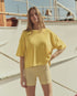 A woman stands outdoors by a boat, wearing The Micro Terry Track Crew by The Great Inc., a loose yellow T-shirt with an oversized silhouette, and yellow striped shorts. Her long light brown hair catches the sun as she looks at the camera on a bright day.