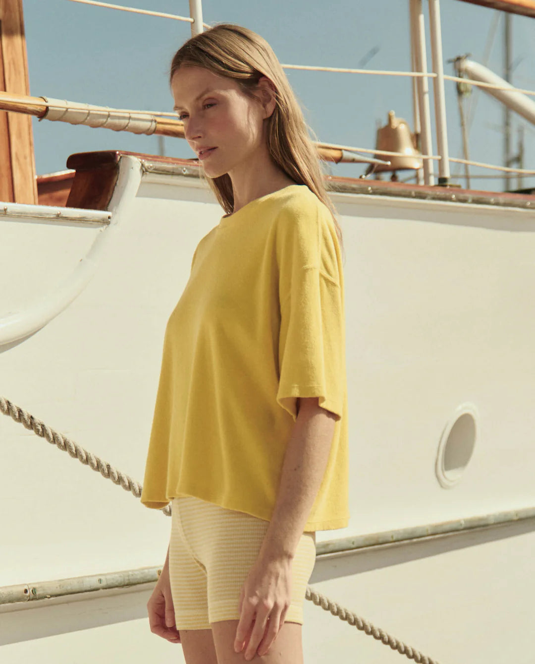A woman with long hair stands outdoors by a white boat under a clear sky, wearing The Great Inc.’s Micro Terry Track Crew—a loose yellow cotton top—paired with light-colored shorts.