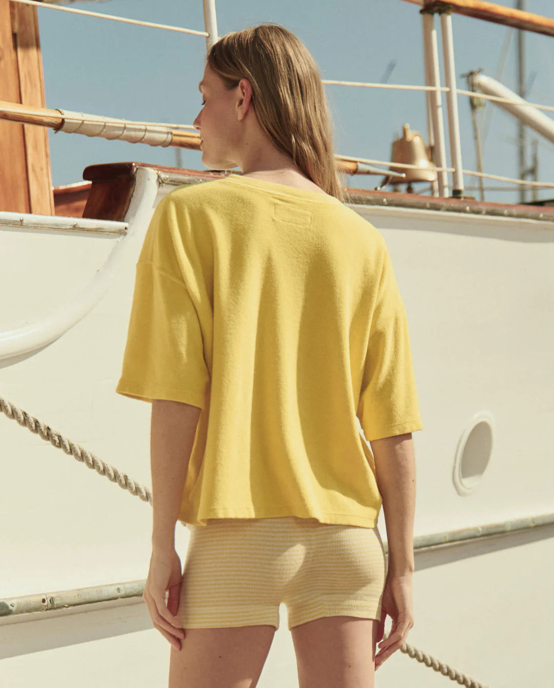 A woman with light brown hair stands on a boat, facing away, wearing The Micro Terry Track Crew by The Great Inc. in yellow and light beige shorts. The sunny sky and parts of the boat’s railing are visible.