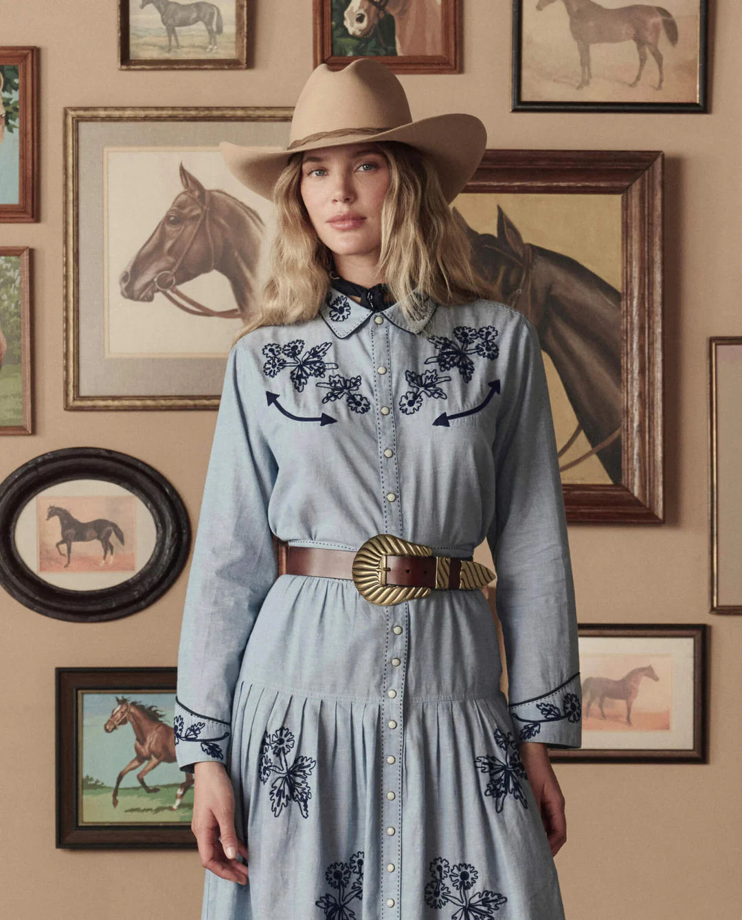 A woman in a tan cowboy hat and blue dress with dark blue embroidery stands before framed horse paintings. She has wavy blonde hair and wears The Great Inc.’s The Big Buckle Belt, featuring Western craftsmanship and a large buckle.