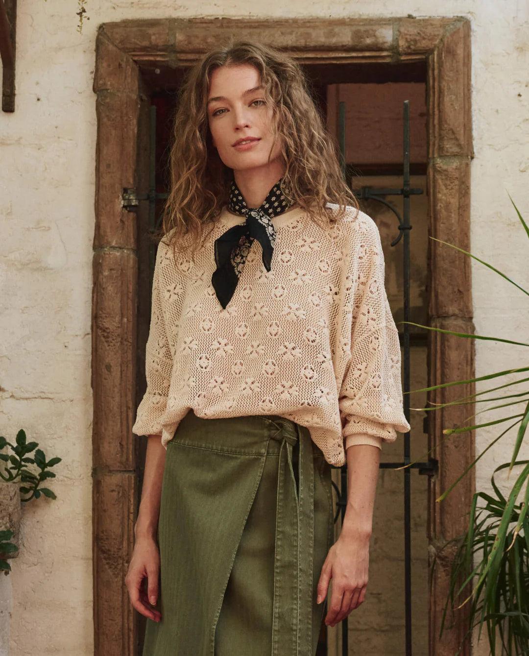 A woman with wavy brown hair stands by a rustic door, wearing The Crochet Pullover by The Great Inc., an olive wrap skirt, and a black scarf with white dots. She looks calmly at the camera.