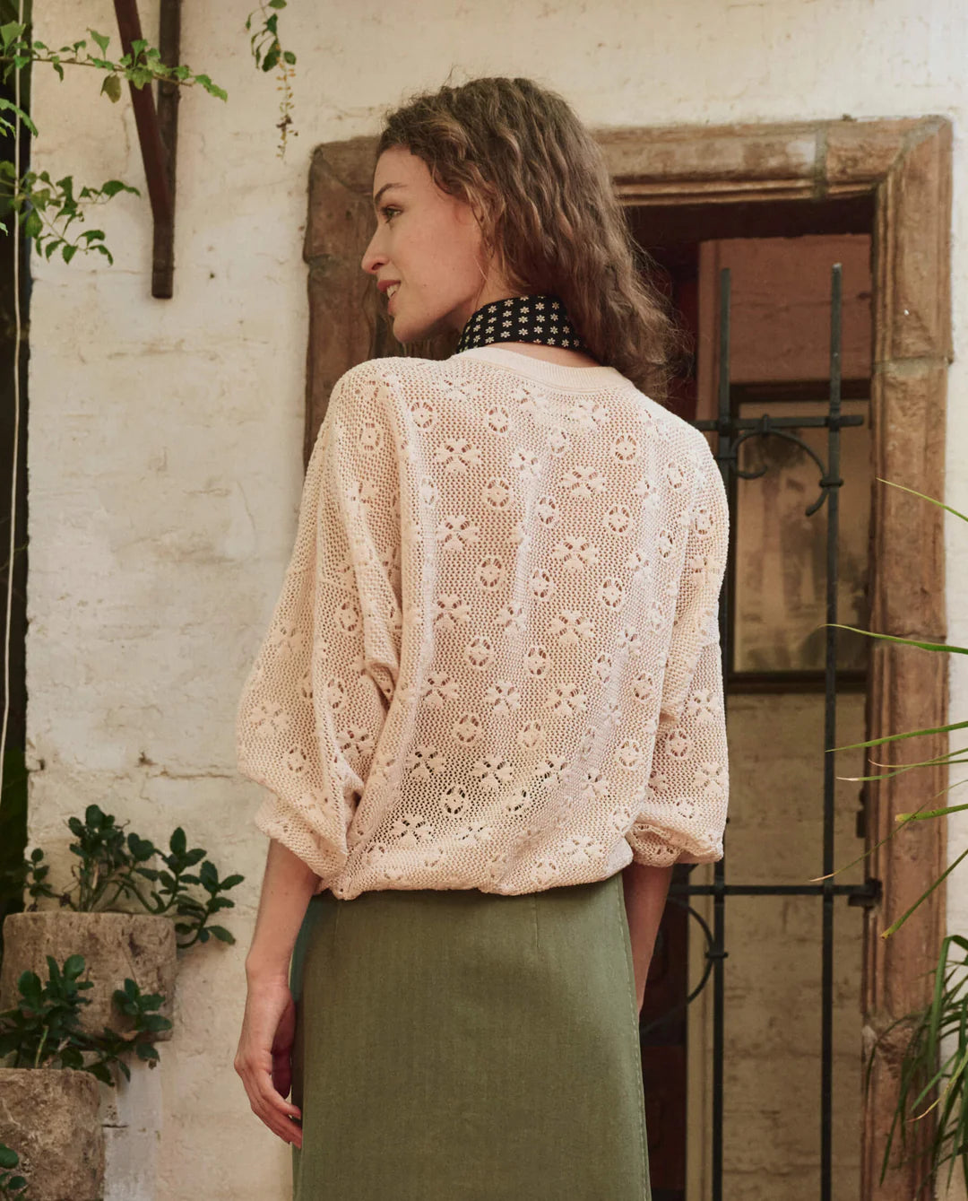 A woman with loose brown waves stands near a rustic stone wall and window, wearing The Great Inc.'s The Crochet Pullover and a green skirt.
