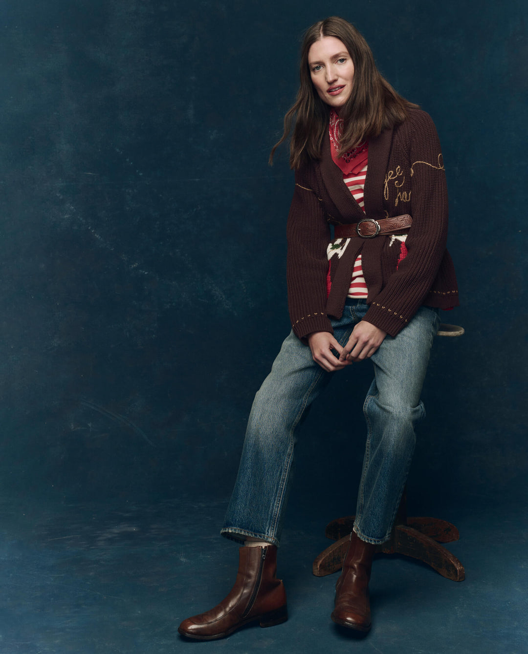A woman with long brown hair sits on a wooden stool, wearing The Great Inc.'s Lasso Lodge Cardigan over a red and white striped top, blue jeans, and brown boots, set against a dark blue studio backdrop.