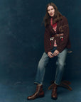 A woman with long brown hair sits on a wooden stool, wearing The Great Inc.'s Lasso Lodge Cardigan over a red and white striped top, blue jeans, and brown boots, set against a dark blue studio backdrop.