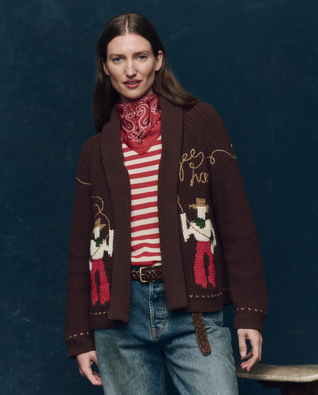 A woman with straight brown hair wears The Great Inc.'s The Lasso Lodge Cardigan with cowboy designs and hand-embroidery, a red bandana, a red and white striped shirt, blue jeans, and a brown belt against a dark background.