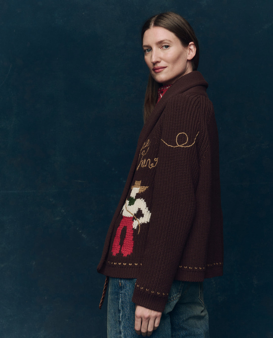A woman with straight brown hair wears The Great Inc.'s The Lasso Lodge Cardigan, its colorful hand-embroidery contrasting her blue jeans as she stands sideways and smiles softly against a dark, textured background.