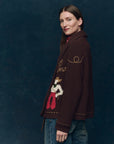 A woman with straight brown hair wears The Great Inc.'s The Lasso Lodge Cardigan, its colorful hand-embroidery contrasting her blue jeans as she stands sideways and smiles softly against a dark, textured background.