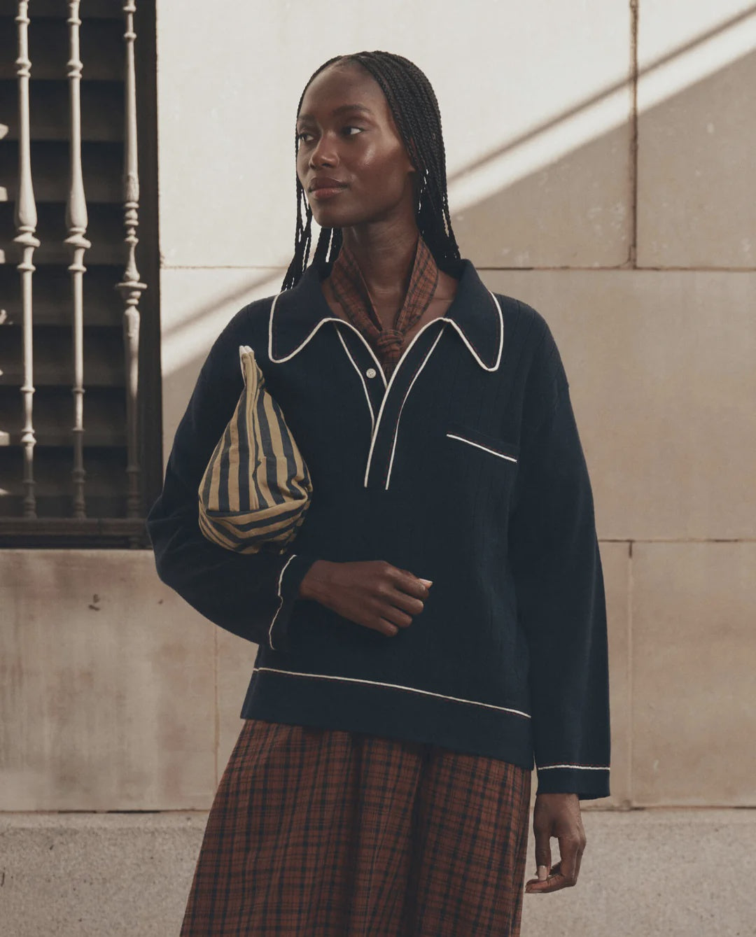 The Great Inc. menswear polo with white trim and a plaid skirt, styled outdoors with a striped bag.