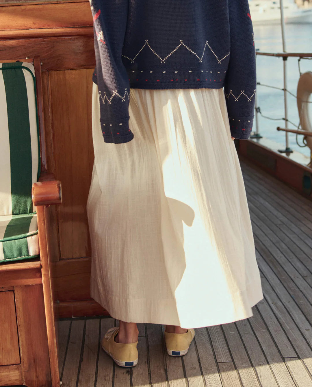 A person stands on a wooden boat deck with their back to the camera, wearing The Viola Skirt by The Great Inc., paired with a navy patterned sweater and tan slip-on shoes.