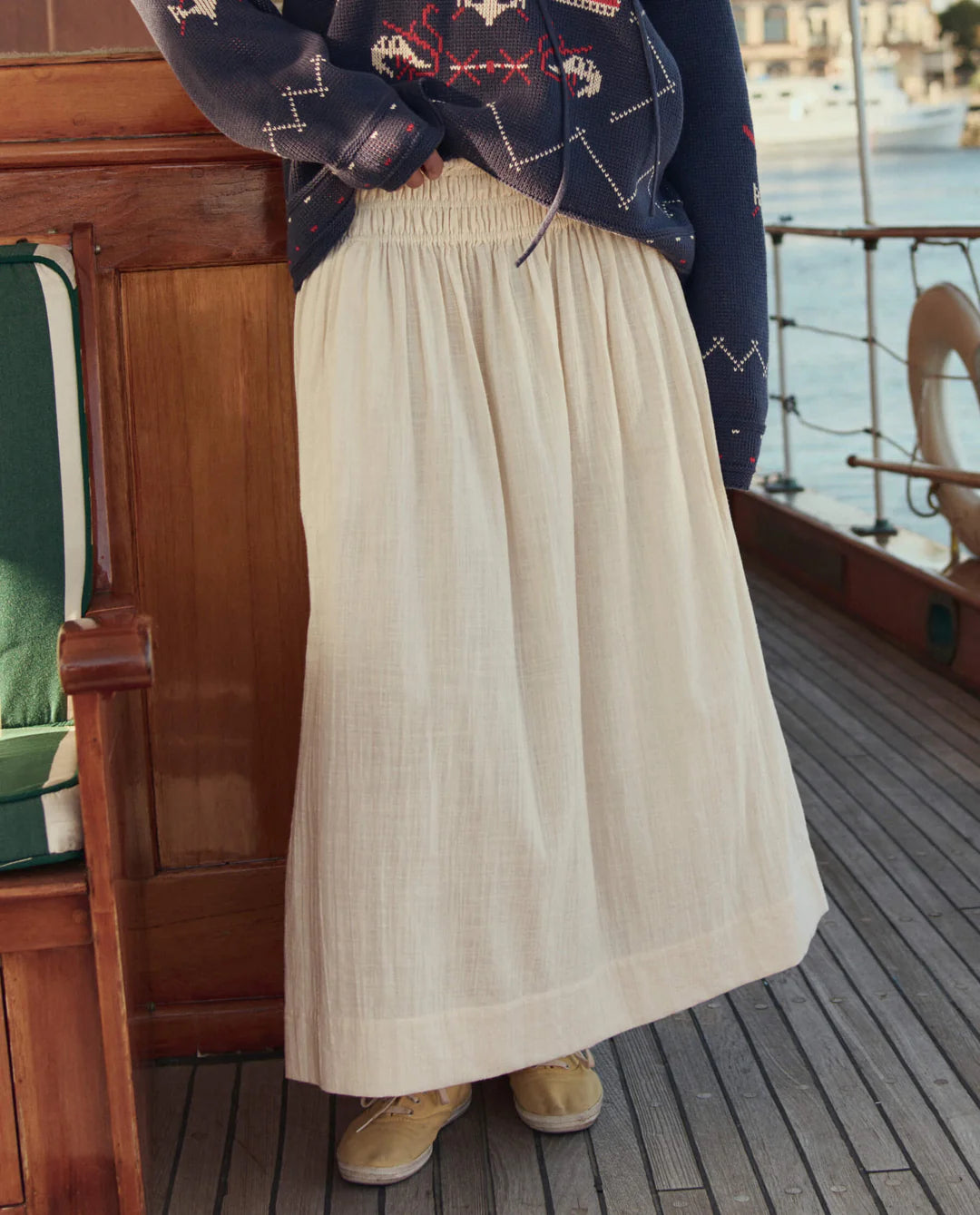 Wearing The Great Inc.'s Viola Skirt—an ankle-length cream cotton skirt with a smocked waistband—a person stands on a wooden boat deck, paired with tan shoes and a navy star-patterned sweater; water and buildings are visible behind.