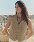 A person with long dark hair stands outdoors in a desert landscape, wearing The Great Inc.'s The Suit Vest in light brown cotton herringbone paired with white pants, mountains and cacti visible in the background.