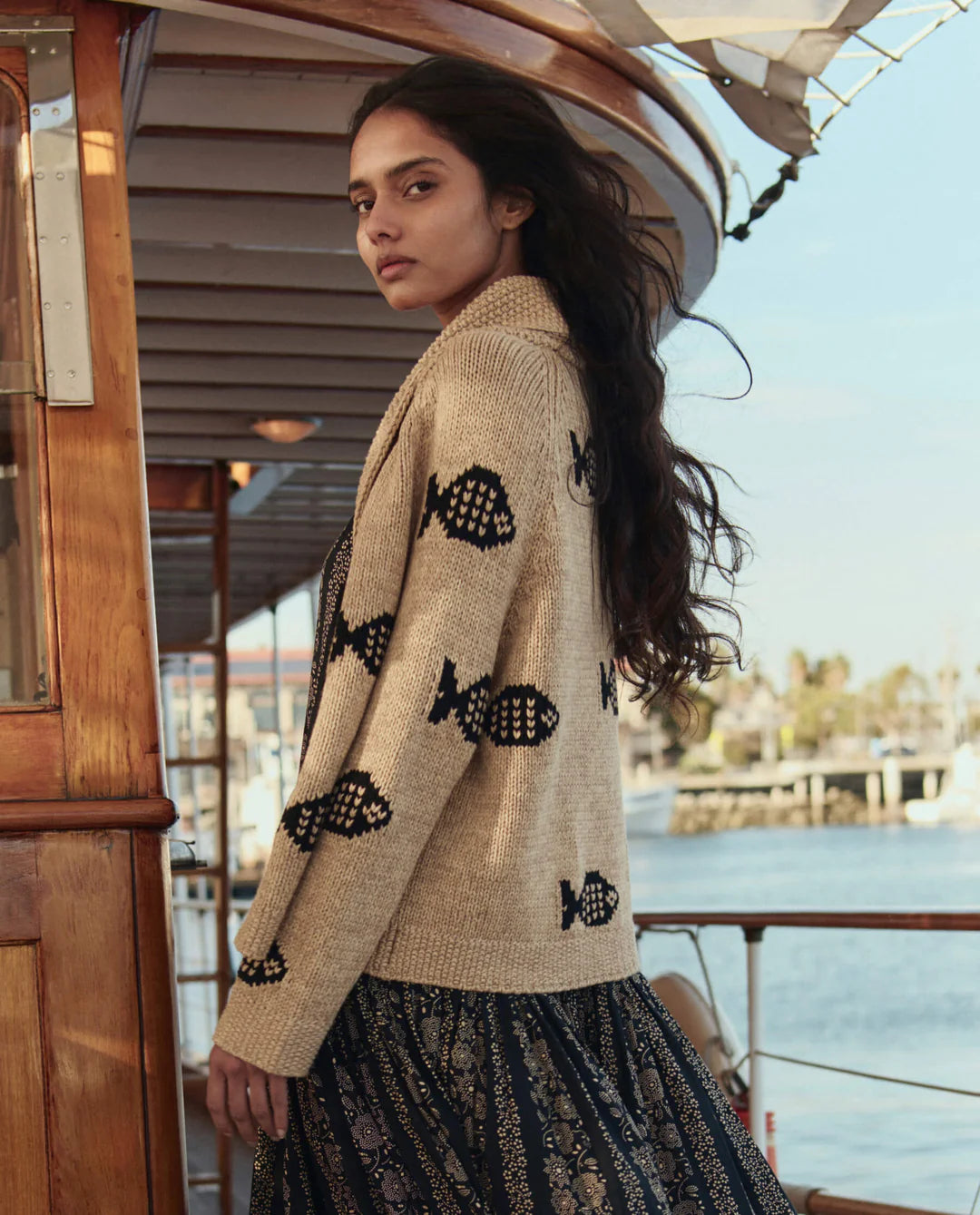 A woman with long dark hair stands on a boat, wearing The Great Inc.'s Swimming Fish Lodge Cardigan featuring a swimming fish motif, paired with a patterned skirt. Water, boats, and a sunny sky fill the background.