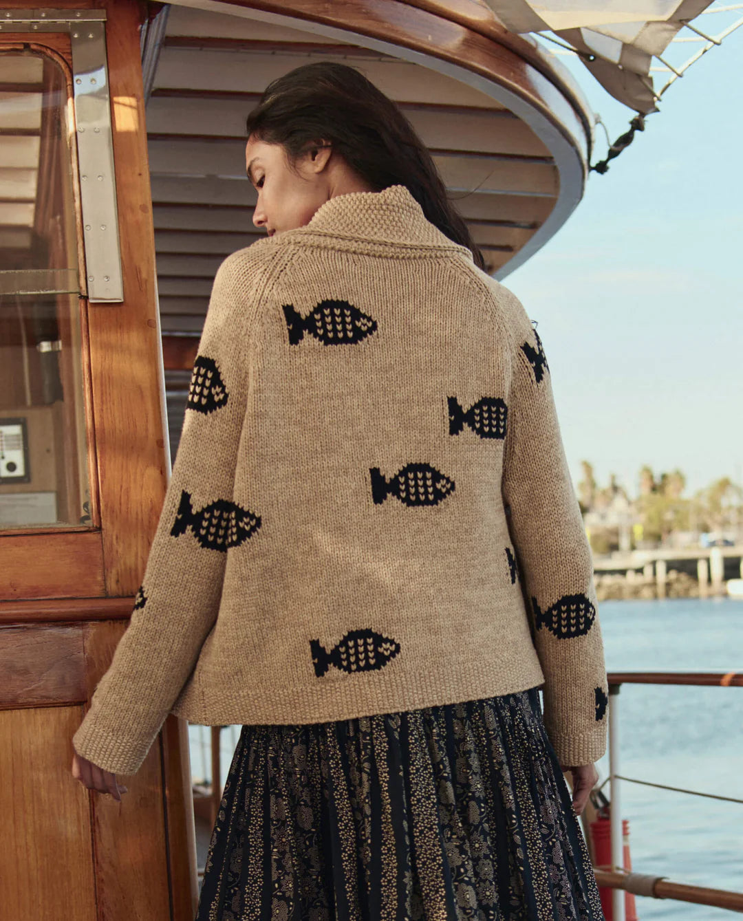 A woman faces away on a wooden boat, wearing The Swimming Fish Lodge Cardigan by The Great Inc., featuring black swimming fish motifs, and a long, dark skirt. Palm trees and water create a serene shoreline backdrop.