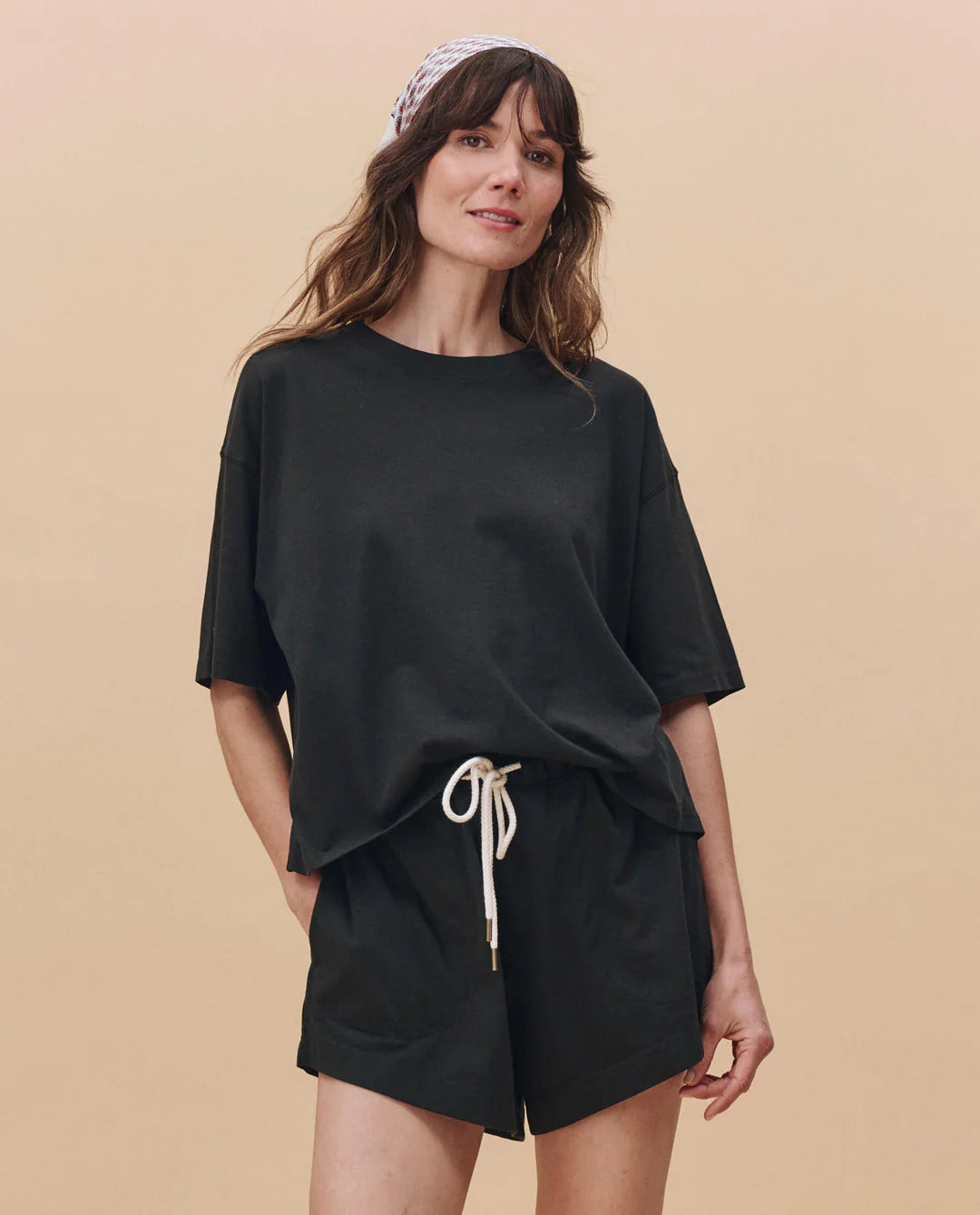 A woman with wavy brown hair and a patterned headband stands against a beige background, wearing The Great Inc.'s black cotton The Flutter Short with a white drawstring and an oversized tee. Her comfy look is made in Los Angeles.