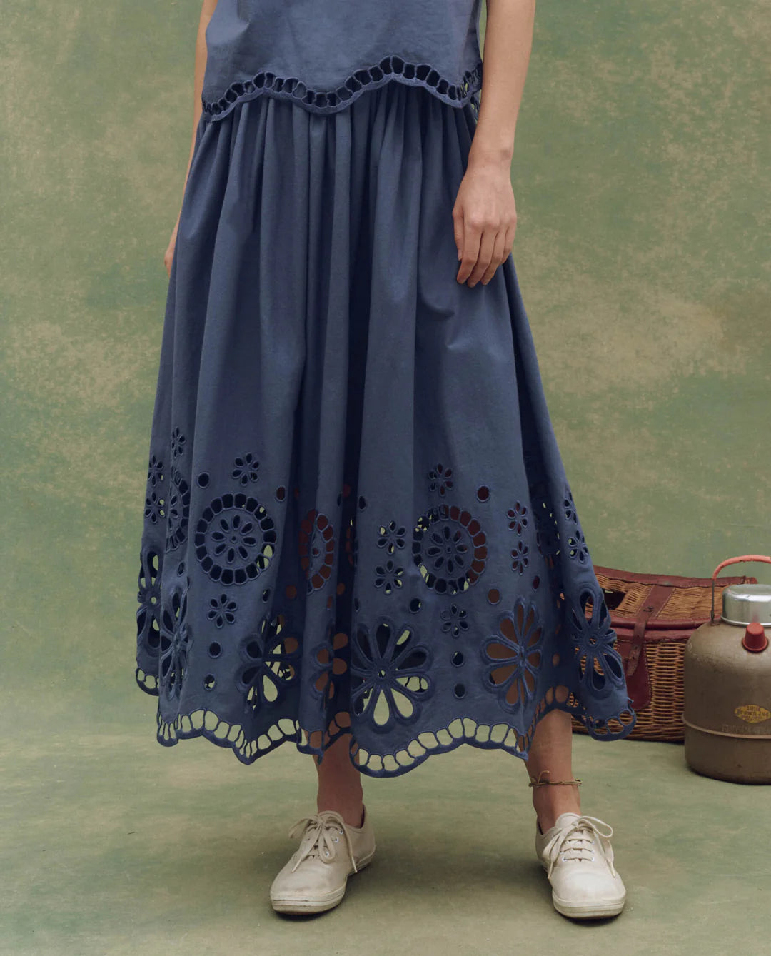 Wearing The Great Inc. Super Bloom Eyelet Skirt with floral eyelet cutouts at the hem, a person stands on a mottled green surface in white shoes, with a vintage picnic basket and metal container behind them.