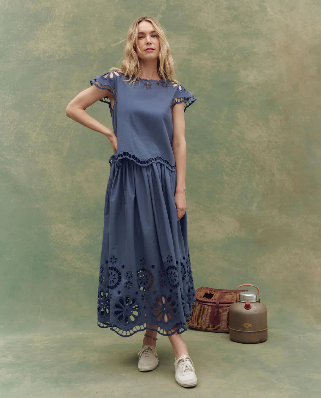 A woman wears The Great Inc. Super Bloom Eyelet Skirt, featuring floral eyelet cutouts, paired with white sneakers. She stands by a vintage basket and metal canister against a green textured backdrop.