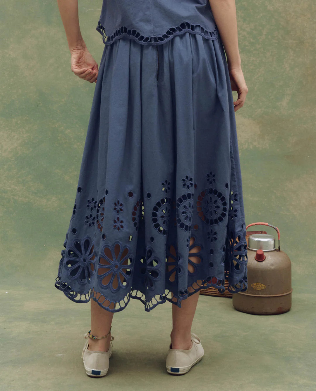 A person stands facing away in The Great Inc. Super Bloom Eyelet Skirt, featuring delicate floral eyelet cutouts at the hem. They're wearing beige shoes, with an old-fashioned kettle nearby for a nostalgic touch.