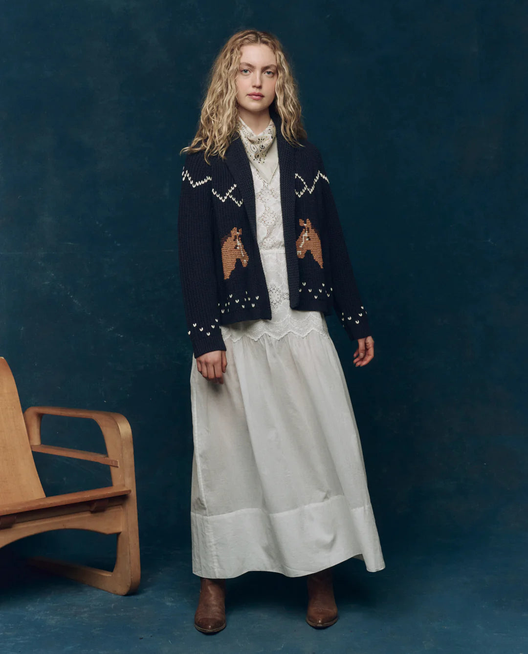 A woman with curly blond hair wears a long white dress, brown boots, and The Great Inc.'s The Horse Lodge Cardigan, featuring hand-embroidered horses, standing by a wooden chair against a dark blue backdrop for western nostalgia.