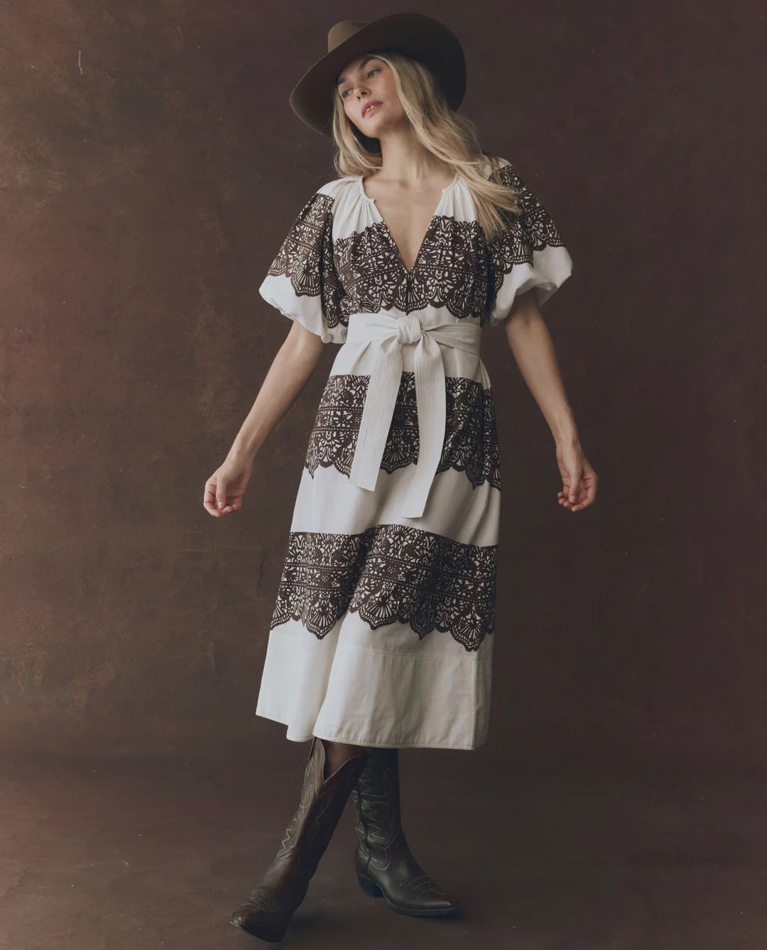 A woman in The Great Inc.'s Fairground Dress with a western lace print, pullover silhouette, wide-brimmed hat, and black cowboy boots poses against a dark brown background.