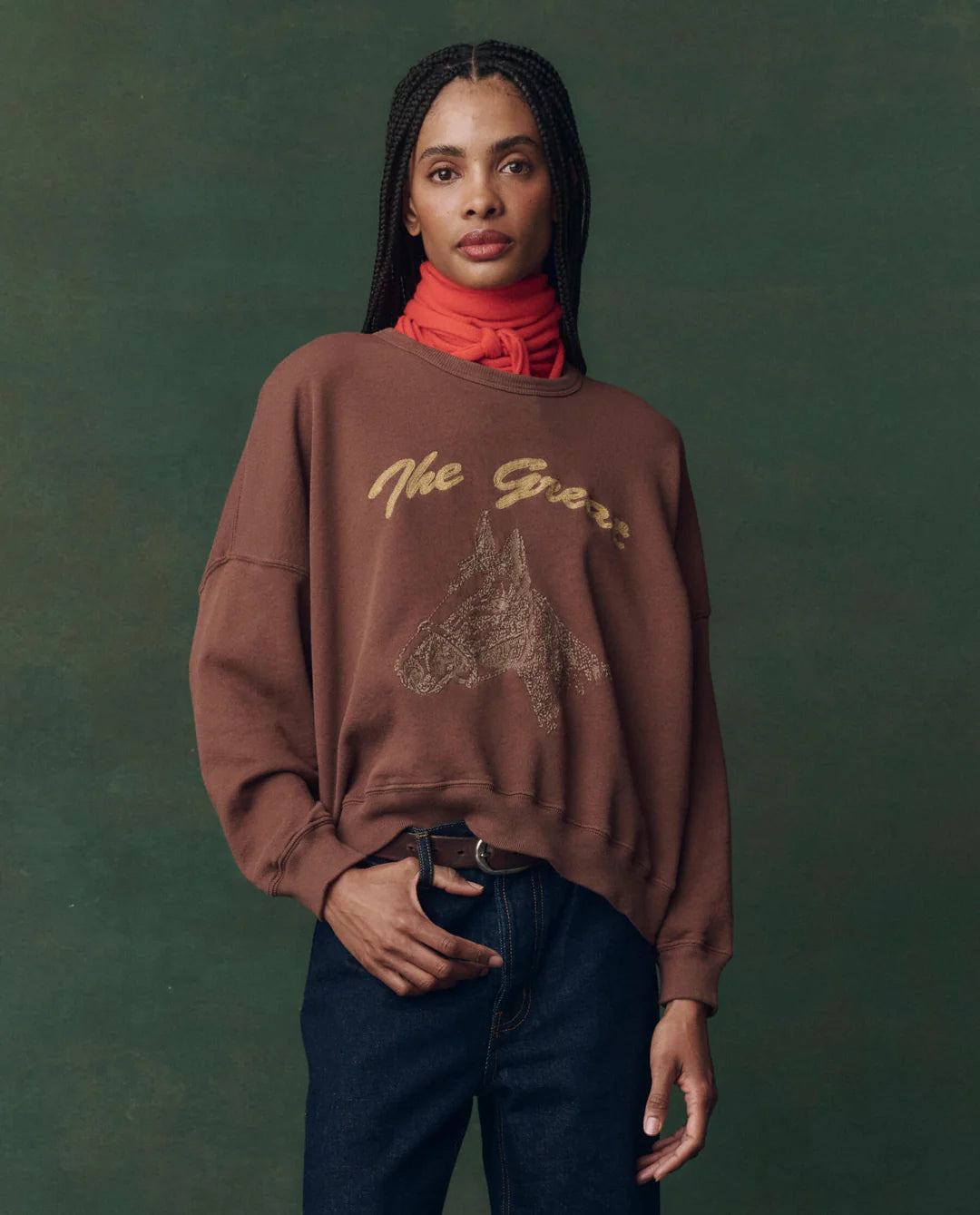 A woman with braided hair wears The Teammate Sweatshirt by The Great Inc., featuring a horse graphic, paired with dark jeans and a red scarf, set against a dark green background.