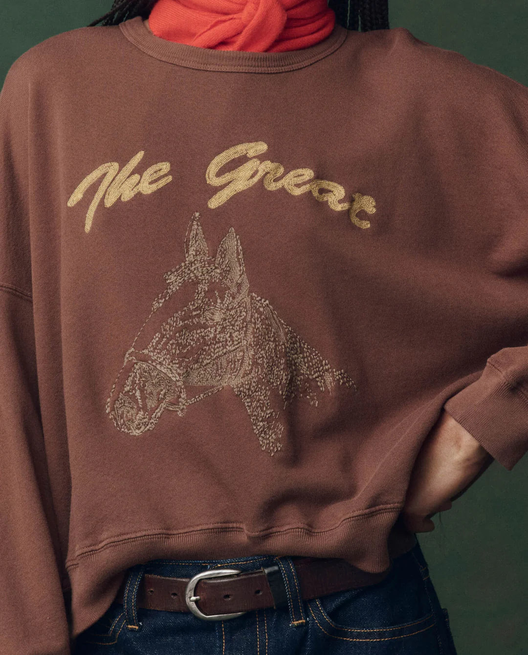 A person wears The Great Inc.'s The Teammate Sweatshirt, a vintage brown top with gold "The Great" lettering and a horse head sketch, made in Los Angeles. They style it with a red scarf, blue jeans, and a brown belt.