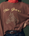 A person wears The Great Inc.'s The Teammate Sweatshirt, a vintage brown top with gold "The Great" lettering and a horse head sketch, made in Los Angeles. They style it with a red scarf, blue jeans, and a brown belt.