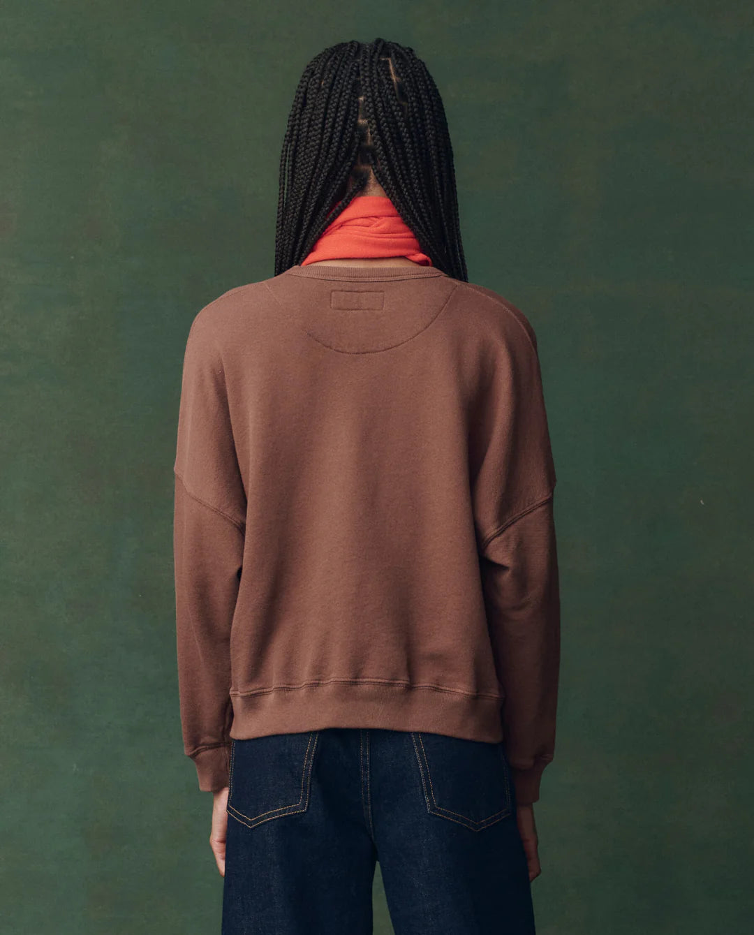 Someone with long, braided hair stands with their back to the camera, wearing The Teammate Sweatshirt by The Great Inc. over a red turtleneck and dark blue jeans, set against a muted green background.