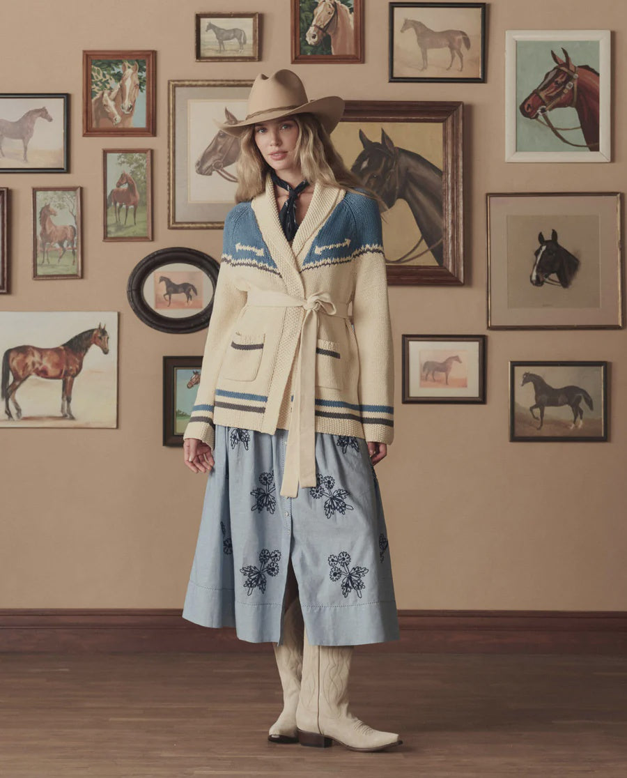 A woman in a cowboy hat, The Western Riding Cardigan by The Great Inc., scarf, and blue floral skirt stands indoors against a beige wall decorated with framed horse paintings and western-themed photos.