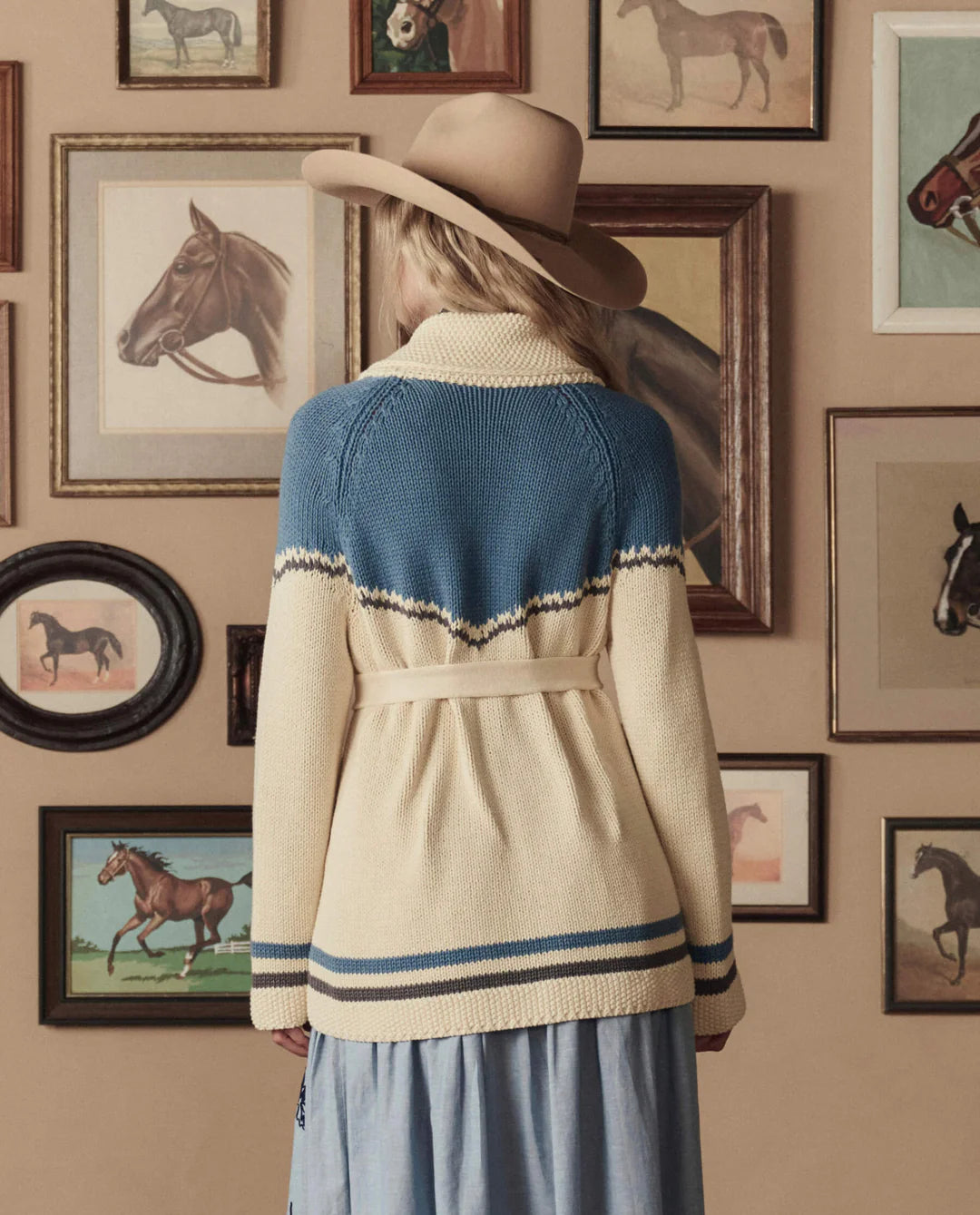 A woman in The Great Inc.'s The Western Riding Cardigan stands with her back to the camera, facing a wall of horse paintings—blending classic Western Americana style with modern fashion.
