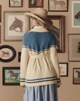 A woman in The Great Inc.'s The Western Riding Cardigan stands with her back to the camera, facing a wall of horse paintings—blending classic Western Americana style with modern fashion.