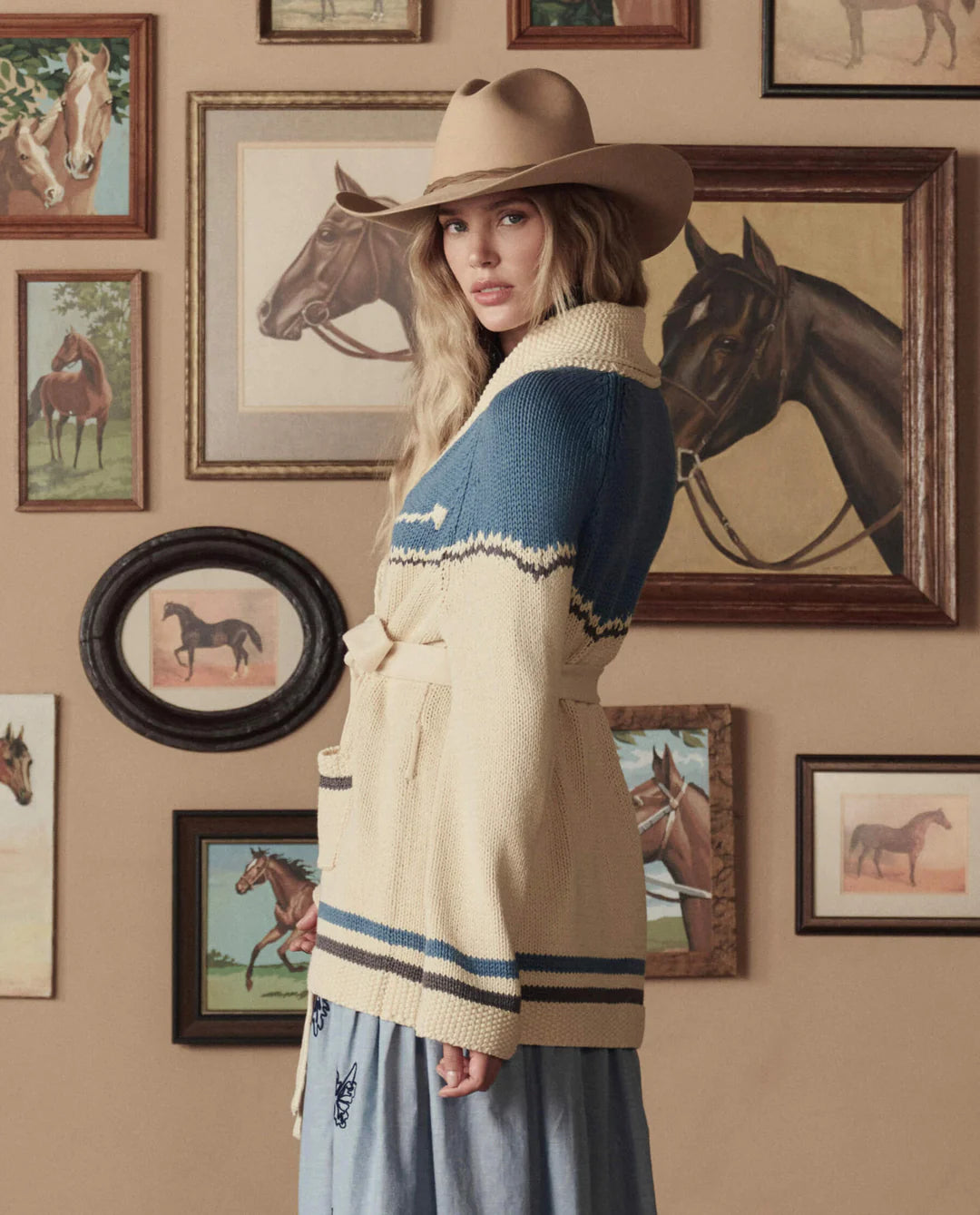 A woman in a tan cowboy hat and The Great Inc.'s Western Riding Cardigan stands before a wall of framed horse art, evoking classic Western Americana as she glances over her shoulder at the camera.