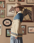 A woman in a tan cowboy hat and The Great Inc.'s Western Riding Cardigan stands before a wall of framed horse art, evoking classic Western Americana as she glances over her shoulder at the camera.