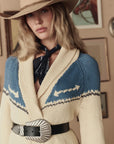 A woman in The Great Inc.'s Western Riding Cardigan in cream and blue, paired with a wide-brimmed hat and silver buckle, stands before framed pictures—showcasing true style from The Great Inc.