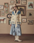 A woman in a cowboy hat, The Western Riding Cardigan by The Great Inc., scarf, and blue floral skirt stands indoors against a beige wall decorated with framed horse paintings and western-themed photos.