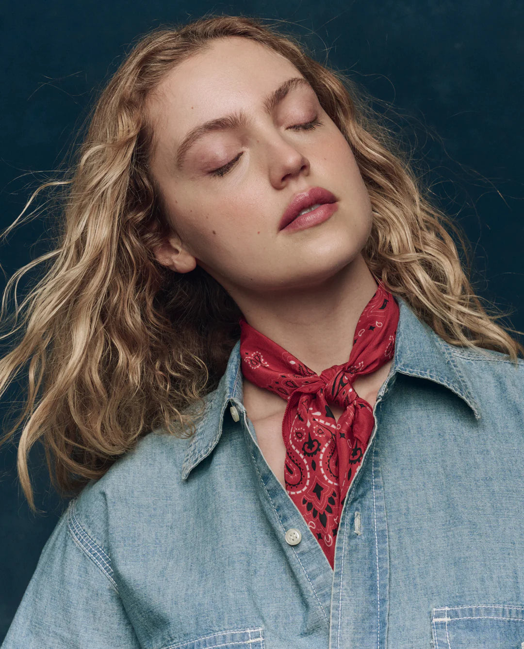 A woman with wavy blonde hair wears a light denim shirt and The Silk Bandana by The Great Inc. around her neck, eyes closed and head tilted against a dark blue background.