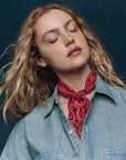 A woman with wavy blonde hair wears a light denim shirt and The Silk Bandana by The Great Inc. around her neck, eyes closed and head tilted against a dark blue background.