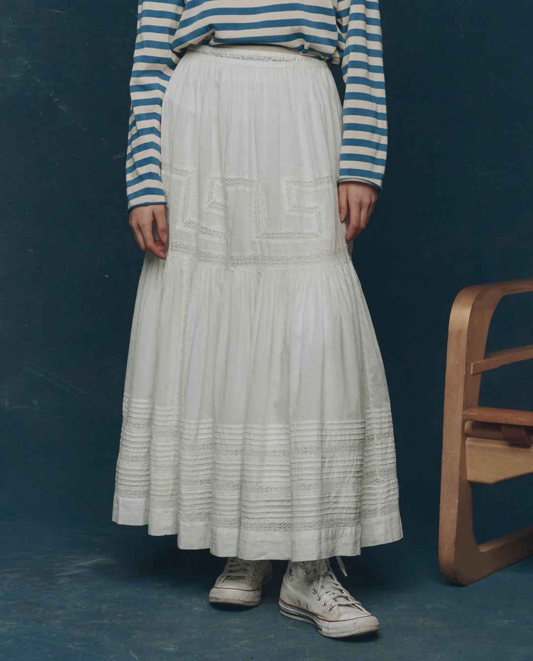 A person wearing a blue-and-white striped long-sleeve shirt, The Great Inc.'s The Victorian Skirt, and white high-top sneakers stands next to a wooden chair against a dark blue background.