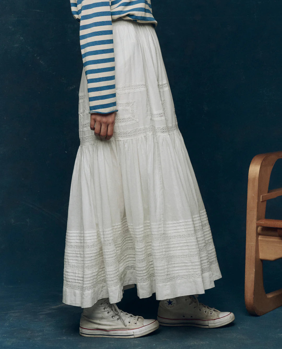 A person in The Victorian Skirt by The Great Inc. pairs it with a blue and white striped long-sleeve shirt and white high-top Converse sneakers, standing beside a wooden chair against a dark blue background.