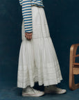 A person in The Victorian Skirt by The Great Inc. pairs it with a blue and white striped long-sleeve shirt and white high-top Converse sneakers, standing beside a wooden chair against a dark blue background.