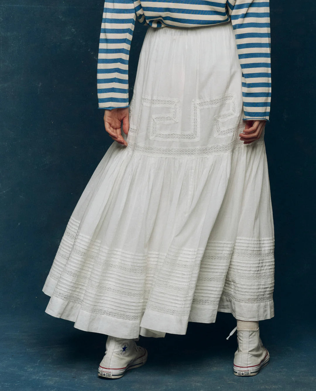 A person wears The Great Inc.'s Victorian Skirt—a long, flowing white lace skirt—paired with a blue-and-white striped long-sleeve shirt and white high-top sneakers, standing against a dark blue background.