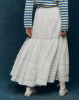 A person wears The Great Inc.'s Victorian Skirt—a long, flowing white lace skirt—paired with a blue-and-white striped long-sleeve shirt and white high-top sneakers, standing against a dark blue background.