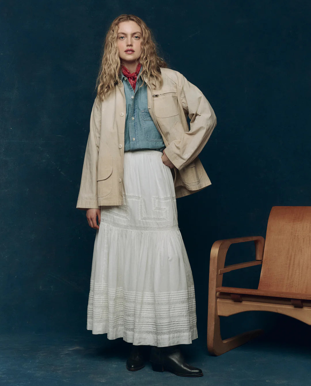 A person stands indoors next to a modern wooden chair against a dark blue backdrop, wearing The Barn Jacket by The Great Inc.—a beige cotton canvas jacket with utility pockets—over a blue denim shirt, white skirt, red neck scarf, and black boots.