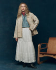 A person stands indoors next to a modern wooden chair against a dark blue backdrop, wearing The Barn Jacket by The Great Inc.—a beige cotton canvas jacket with utility pockets—over a blue denim shirt, white skirt, red neck scarf, and black boots.