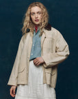 A woman with wavy blonde hair wears The Barn Jacket by The Great Inc., made from garment-dyed cotton canvas, over a denim shirt and a white lace-detailed skirt, standing before a dark blue background.