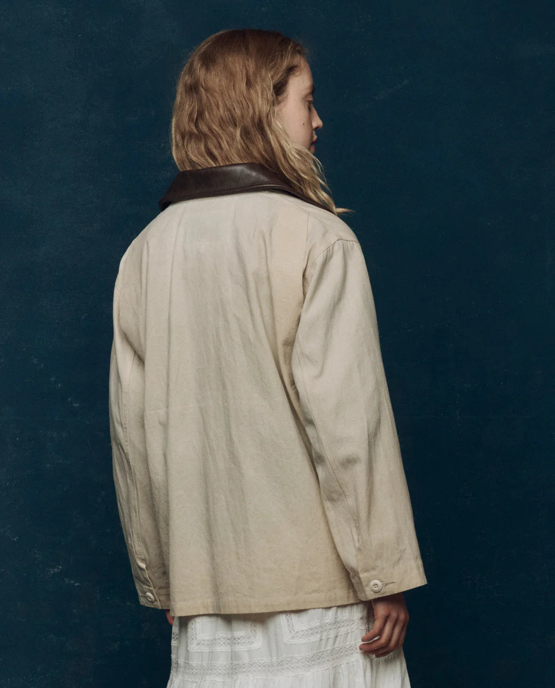 A person with wavy, light brown hair is seen from behind wearing The Great Inc.’s cream-colored Barn Jacket with a dark collar over a white dress, standing against a dark teal background.