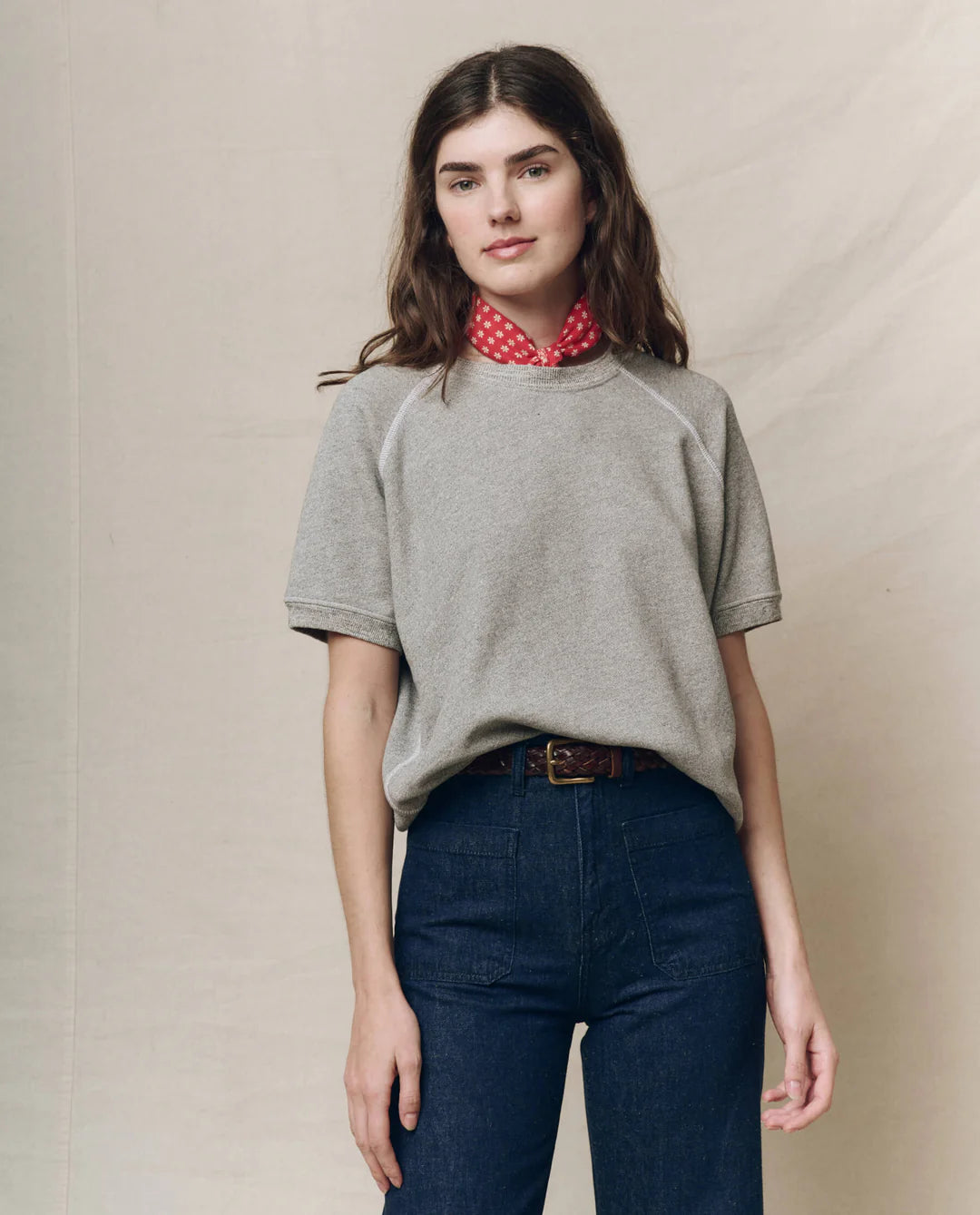 A person with long brown hair stands against a neutral background in The Great Inc.'s The Short Sleeve Sweatshirt, featuring a wild panther graphic. Layered over a red polka dot collared shirt and paired with high-waisted blue jeans, they pose relaxedly with a subtle smile.